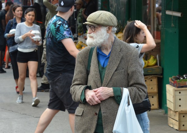 an older man in tweed jacket and cap with long white beard and mustache, black rim glasses, white plastic bag over one arm, walks down the street