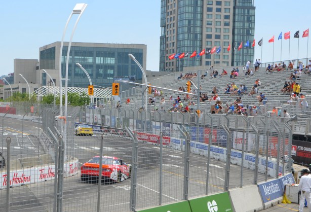 two nascar type cars racing on a track at the Honda Indy, some people sitting in the stands, 