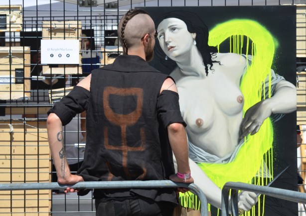 a man, with back to the camera, stands beside a painting of a topless woman, their faces are close together 