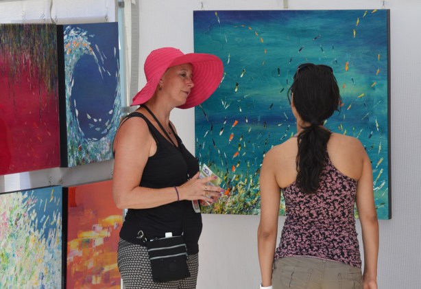 a woman in a large brimmed pink hat is talking to another woman in front of some paintings at an outdoor art fair 