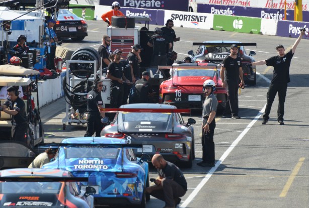 5 Porsche racing cars in the pit lane with many people around them, at Honda Indy in Toronto