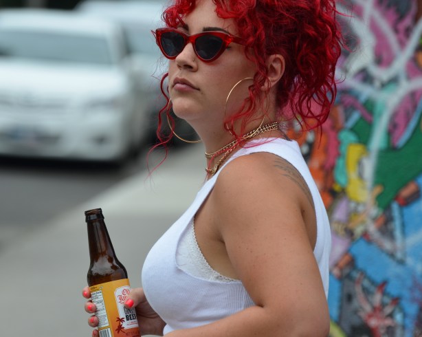 a woman in white sleeveless top and with very red dyed very curly hair, carrying a bottle of ginger beer, 