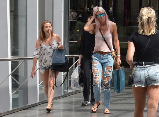 people walking across pedestrian bridge between the Eaton Centre and the Bay, one woman has long light pink hair and is wearing jeans with many rips and holes in the front, 