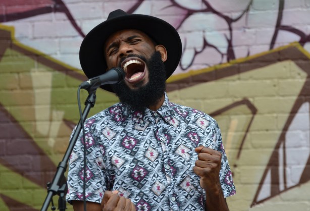 a young black man in black wide brimmed hat sings in front of a microphone, part of a band performing ouside in front of a mural on a wall 