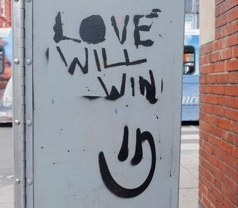 on the side of a grey metal box, written in black, with a smiley face under the words, love will win
