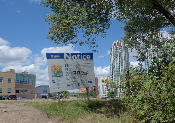 blue and white city of Toronto development notice sign on a small hill, by some trees, in front of a vacant lot. Highrises in the background