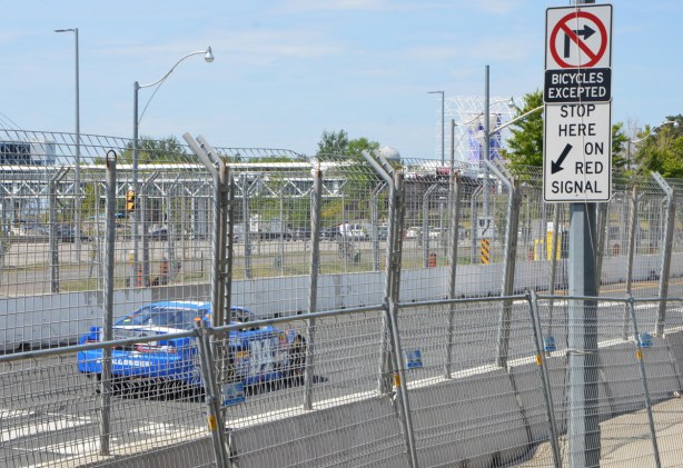 a blue car races on track with is along Lakeshore, behind two layers of chainlink fence 