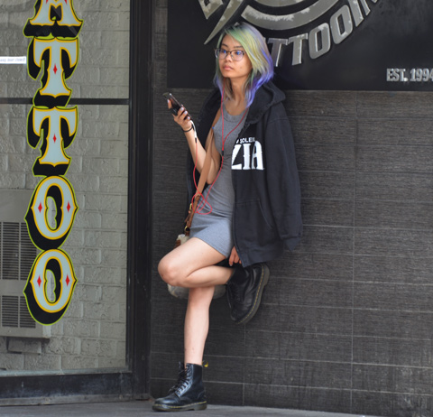 a woman standa against an exterior wall, beside a tottoo place. The word tattoo is in large yellow letters beside her, her hair is multicoloured, she is wearing a short dress, knee bent with one foot against the wall, phone in hand, black jacket over her shoulders 