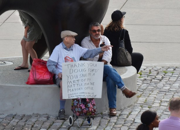 people sitting by the Archer, a Henry Moore sculpture, at Nathan Phillips Square. An older man is talking to another man. He is holding a hand written sign that says Thanks to Doug Ford 