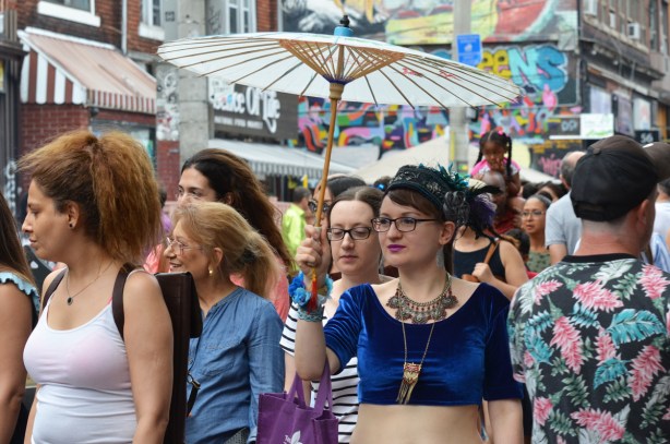 woman in short velvet close fitting top, large necklace, and holding a white parasol walks down a street in a crowd of people