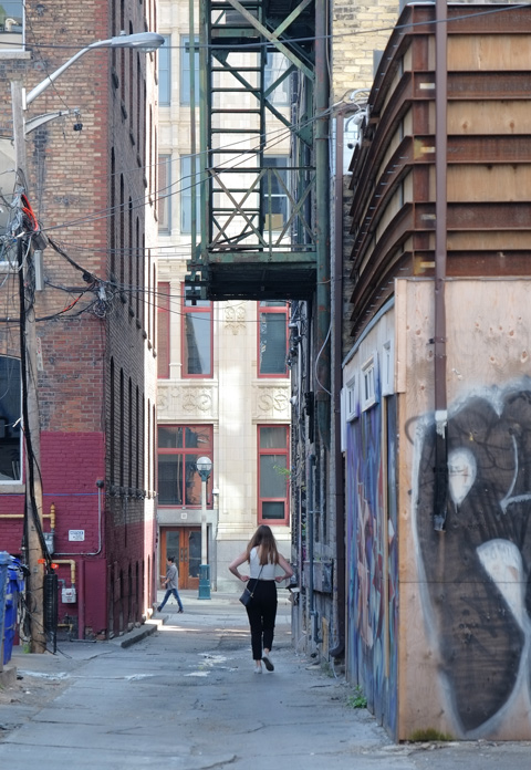 a woman walks down an alley, away from the camera, metal fire escape staircase is above her, brick buildings beside her 