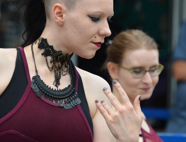 two women dancing, one with long hair on the top but shaved on the sides, wearing an intricate black necklace, double, one woman in focus in the front, the other slightly out of focus, purple and black sleeveless tops, close up of head and shoulders and one hand with grey fingernails