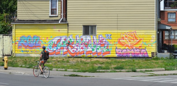 a man cycles past a pale yellow house on Dundas St that has has a bright coloured mural across the bottom of it, text, in blues and pinks 