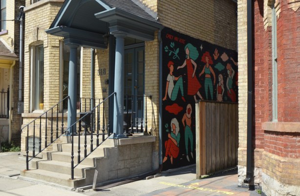 a mural by Emily May Rose of figures of woman on a black background on the side of a building, a fence runs parallel to it, entrance ways to buildings 