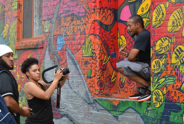 graffiti alley, a group is shooting a music video, black man in on window sill lip synching the words while a woman films 