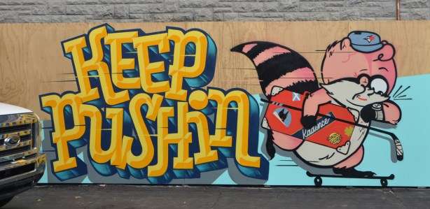 large yellow letters that say keep Pushin, on a mural with a pink raccoon wearing a blue jays baseball cap, looking at a wrist watch with a dollar sign on the face, 