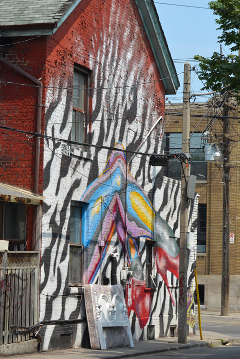 an old mattress and white head board lean against the wall, outside, of a house that has a large mural painted on it of legs with feet in bright red high heeled shoes 