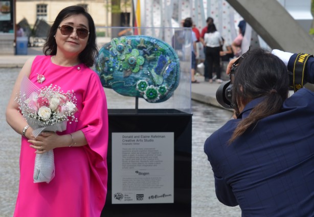 woman in a bright pink dress, holding a bouquet of flowers poses beside a brain artwork at Nathan Phillips Square while a man in a blue suit takes her picture