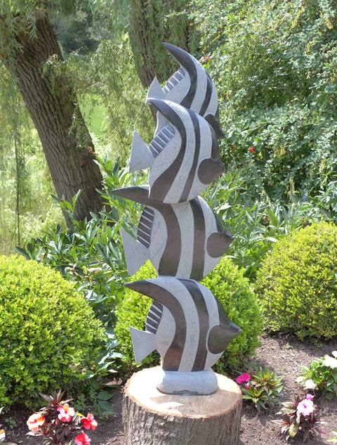 a sculpture titled Shoal, 4 striped fish, one on top of the other, sculpture with flowers in front and greenery behind, Edwards Gardens 