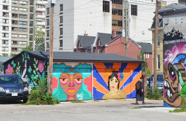 An alley way with a double garage and a car parked beside it, high rise buildings behind it, a young man walking past it. Each door is painted with a mural of a woman's head, one by Desire Betty (dbetty13) and the other by Sarah Gilmore, or toest. On the side of the garage is another mural, this one by nightarcade, Sarah Cannon, 