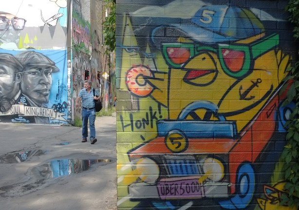 A man avoids the puddles in the lane as he walks past an uber5000 mural of a yellow birdie with a donut, birdie is driving a little orange car 