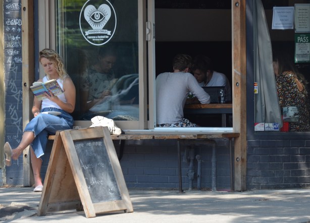 a woman sits outside a coffee shop reading a book. the window of the coffee shop is open and there are 4 people sitting inside 