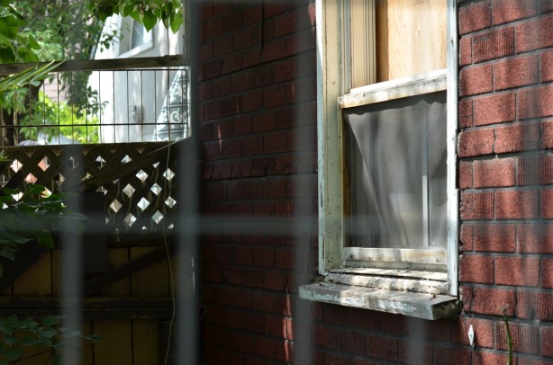 behind a metal fence, an old house with a boarded up window 