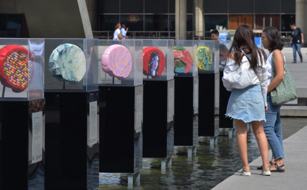 two young woman are standing beside a row of artwork, brain shaped, each in a plexiglass container, standing in the water of the fountain at Nathan Phillips Square, 7 brain sculptures, all decorated by different artists, part of the brain project in support of Baycrest
