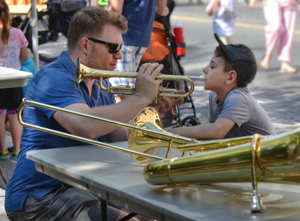 a man teaches a boy to blow a trombone, outdoors, activity on the sidewalk during Open Streets 