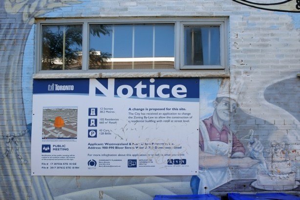 blue and white city of toronto development notice sign on the side of a building, under a window, beside a mural of a baker in chefs hat and white apron icing a three layer wedding cake 
