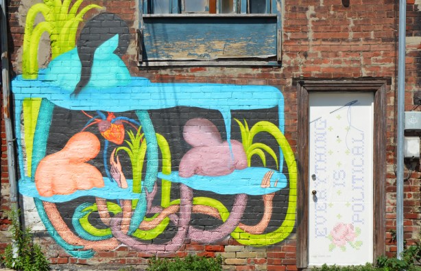 a white door with words that say Everything is political, also a large mural painted beside the door, two figures amongst pink, yellow and pale blue curly vines and leaves, abtract 