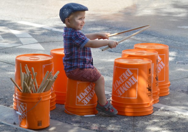 a young boy uses wooden drum sticks to bang on upturned orange plastic buckets from Home Depot, outside, activity at Open Streets