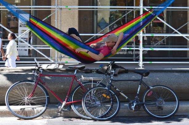 a young couple share a hammock strung between two trees on a diving strip own the middle of Yonge Street, their bikes are parked beside the hammock