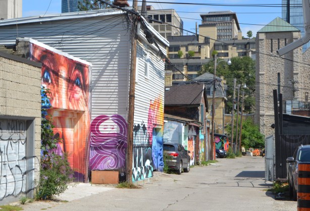 laneway in toronto with some garages with murals on them, tall buildings in background, utility poles and wires, 