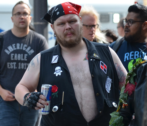 man in black and red bandana, black vest open to reveal bare chest, tattoo above his heart, black glove, carrying a can of red bull