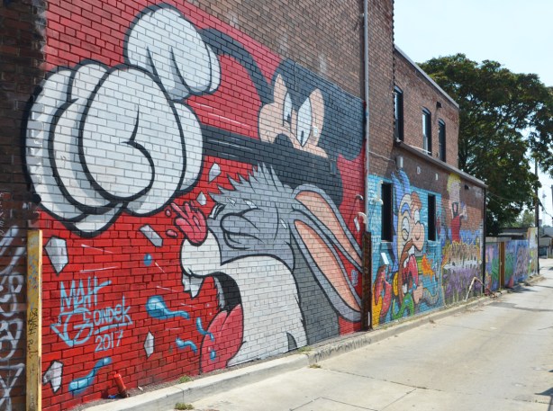  in a popart mural by Matt Gondek and Jackson, in an alley, mickey mouse with his big white gloves is punching bugs bunny whose mouth is wide open and tongue is out 