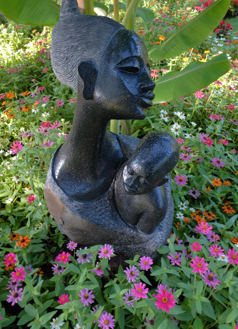 black stone sculpture of a mother holding her son, in a garden, with pink, red and orange flowers around the bottom of the sculpture