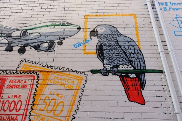 part of a mural in a narrow alley in Kensington by Dave Setrakian, a parrot on a perch and an airplane, some passport stamps