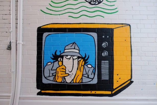 part of a mural in a narrow alley in Kensington by Dave Setrakian, a parrot on a perch and an airplane, a television set showing Ispector Gadget, a cartoon character from a 1990's kids program