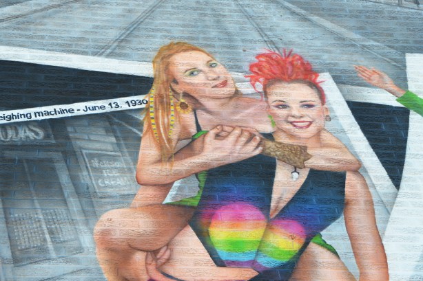 painting of two young women, one blond and one withshort curly bright red hair, on top of painting of black and white vintage photos 
