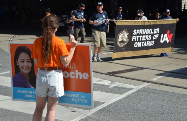 labour Day parade on Queen St West, a young woman holds an orange NDP sign for the local Parkdale MPP Bhutial Krapoche, as members of the Sprinkler Fitters of Ontario Union come around the corner from Queen West to Dufferin 