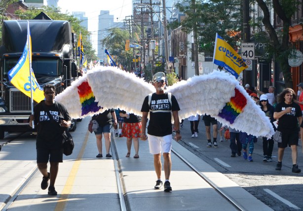 labour Day parade on Queen St West, a man walks with large white wings, about 8 feet across (2.5 metres), he's wearing a black T-shirt with white words that say No Justice No Peace 