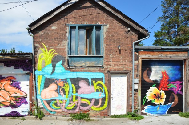 old two storey brick building in a lane with upper window and lower single door. Door has a street art piece on it as does whole lower level, a garage on either side of it each with the door covered in a new mural 