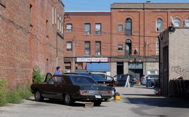 man stands beside on older car in a parking lot surrounded by old brick buildings