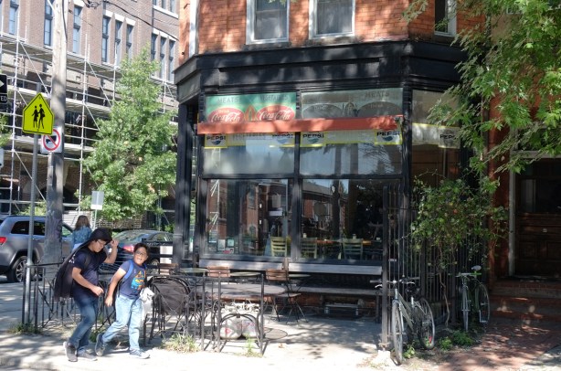 a deli, cafe, with old coca cola, coke, signs as well as pepsi signs. chairs and tables out front, large windows, two boys wakling past, on a corner in a residential area, old house