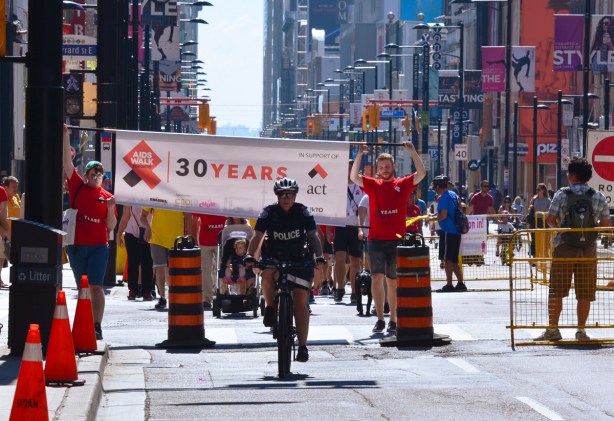 a policeman on a bike leads an AIDS walk up Yonge Street, people holding a banner follow him and then many people wearing red T-shirts