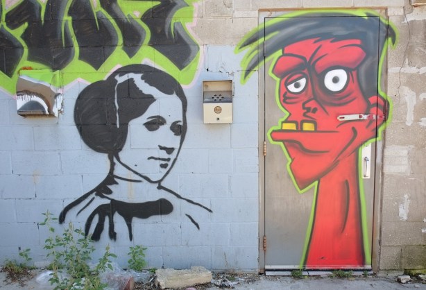 street art in an alley - red man's head, with protruding lower jaw and two large yellow teeth, white eyes, on a door, black and white picture of Star Wars Princess Leia on the wall beside 