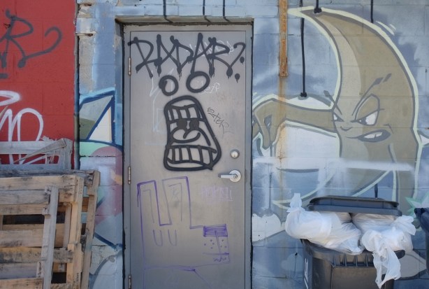 street art in an alley - word radar on grey metal door, with screaming face below, on the wall beside is a moon shpaed figure, with arm out and seems to be holding something in its fingers but nothing there, garbage bins (real) below 