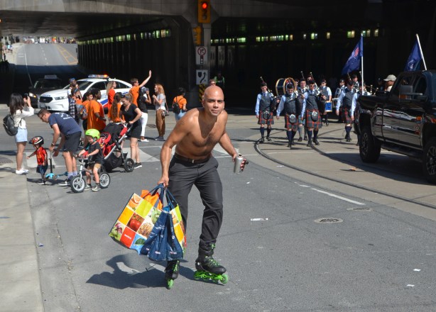 a man roller blades past the parade, water bottle in one hand, grocery bags in the other, he is topless, labour Day parade on Queen St West, 