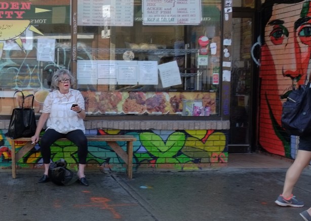 a woman in a white blouse sits on a bench ooutside a restaurant, on a wood bench, smoking a cigarette 